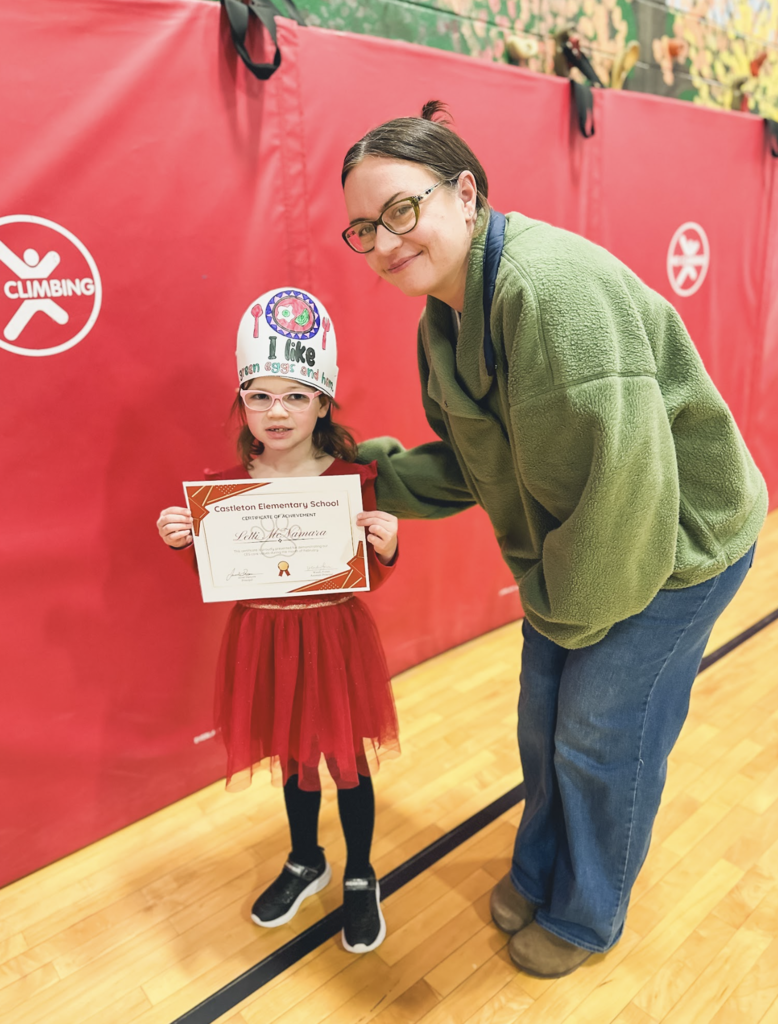 A young student wearing a paper crown stands with a staff member while holding a Castleton Elementary School certificate of achievement.