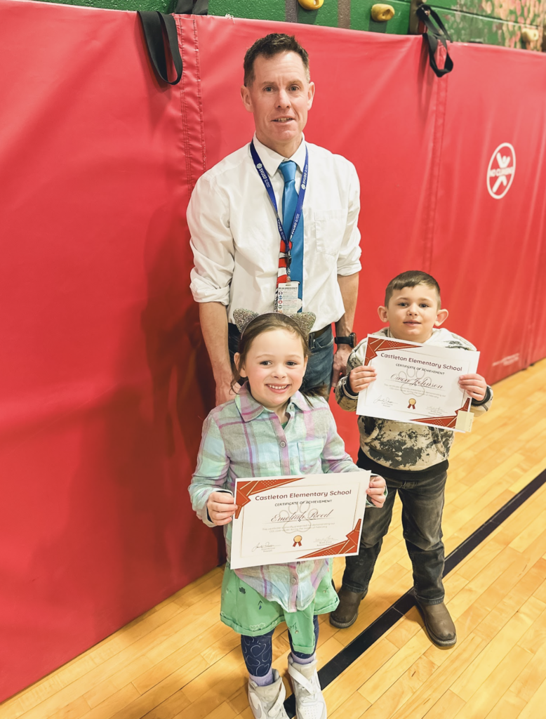 Two elementary students stand in a school gym holding Castleton Elementary School certificates of achievement beside a staff member.