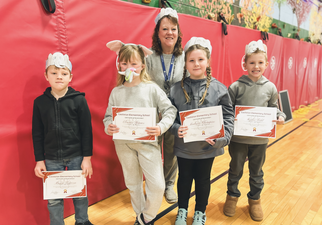 Four elementary students wearing animal-themed paper headbands stand with a teacher in a gym, each holding a Castleton Elementary School certificate of achievement.