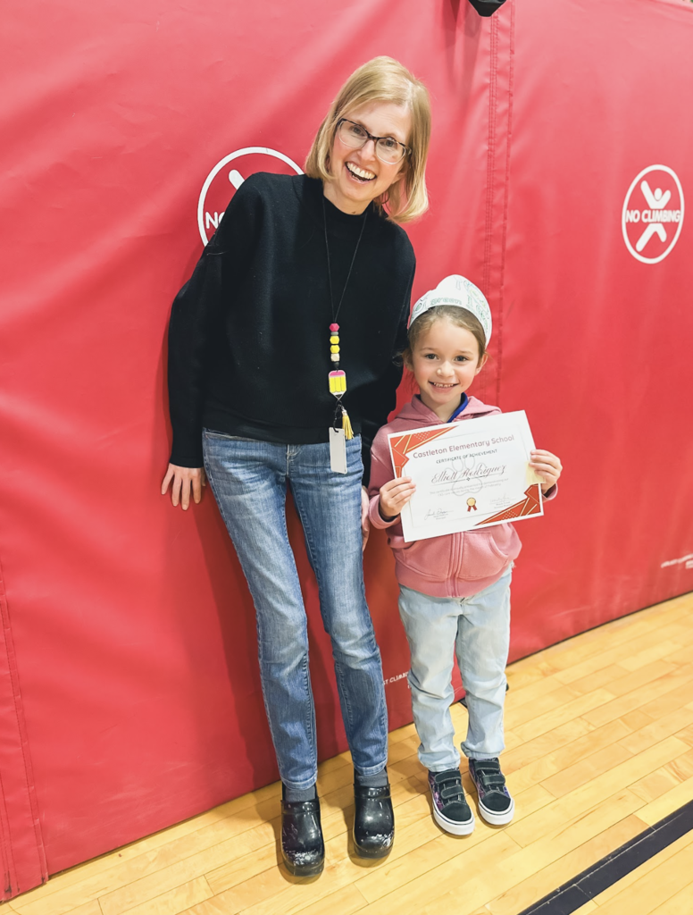 A teacher and elementary student stand together in a gym while the student holds a Castleton Elementary School certificate of achievement.