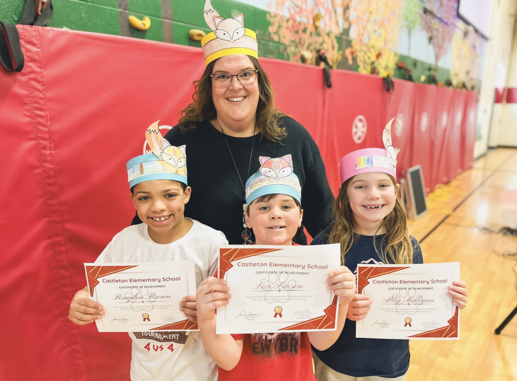 Two students wearing fox-themed paper headbands stand with a teacher in a gym while holding Castleton Elementary School certificates of achievement.