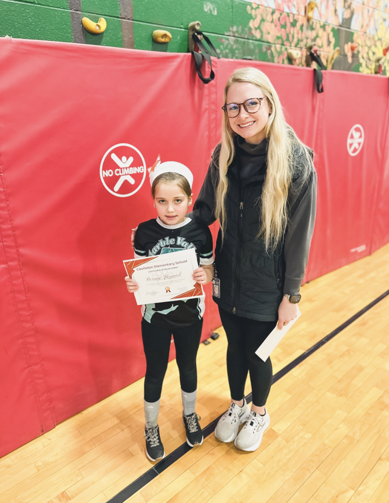 Young student holding a Castleton Elementary School certificate stands beside a staff member in the gym.