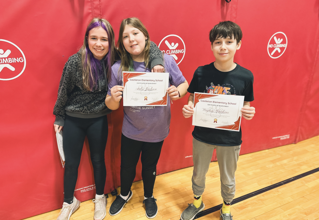 Three elementary students stand together in the gym holding Castleton Elementary School certificates of achievement.