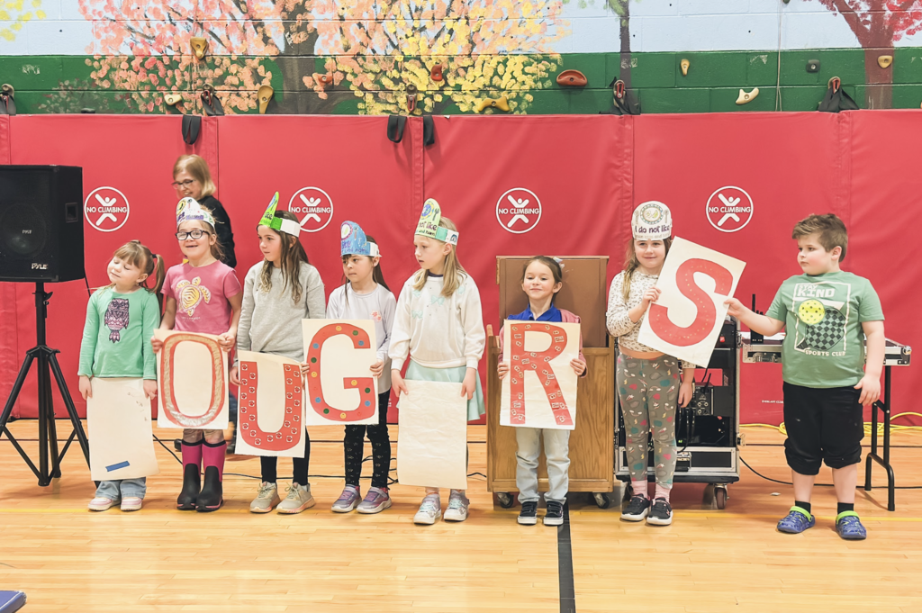 Group of young students stand in a line in the gym holding large letters that spell “COUGARS.”
