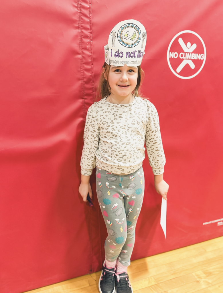 Student wearing a paper crown reading “I do not like green eggs and ham” stands in front of red gym mats.