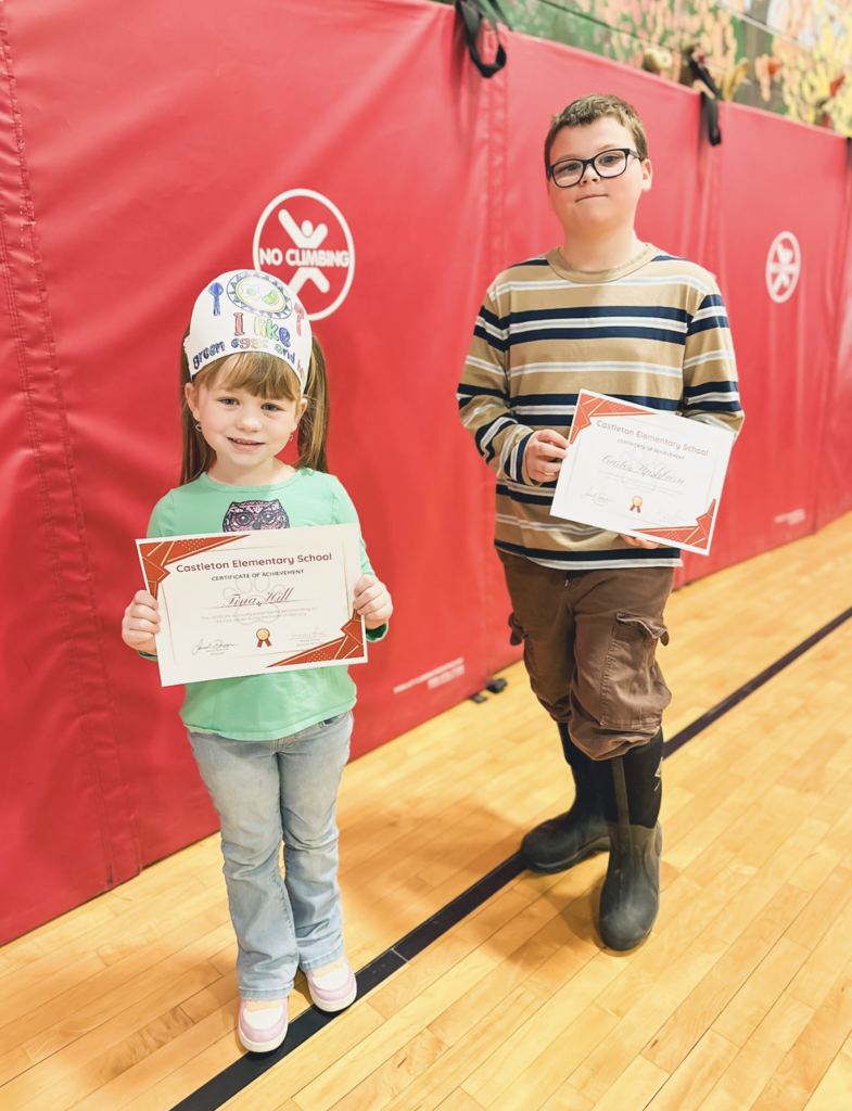Two elementary students walk in the gym holding Castleton Elementary School certificates of achievement.