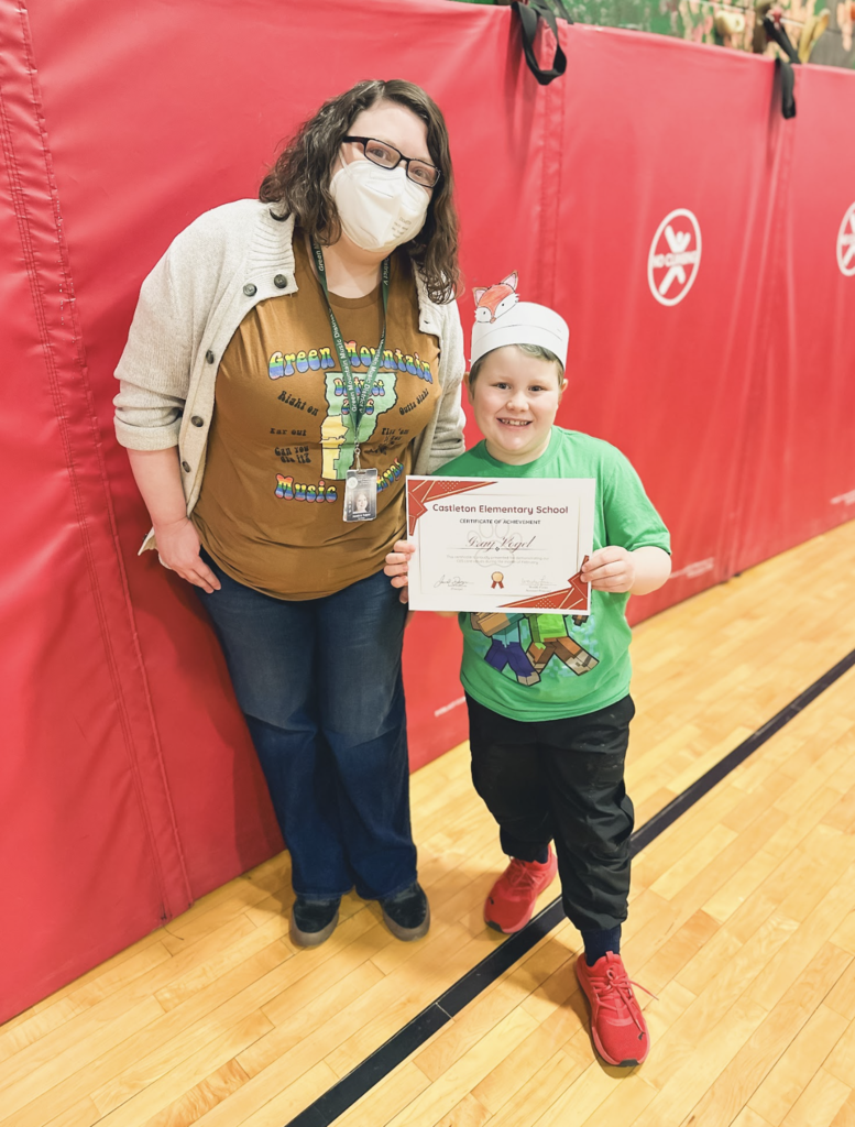 Student holding a Castleton Elementary School certificate stands beside a staff member wearing a mask in the gym.