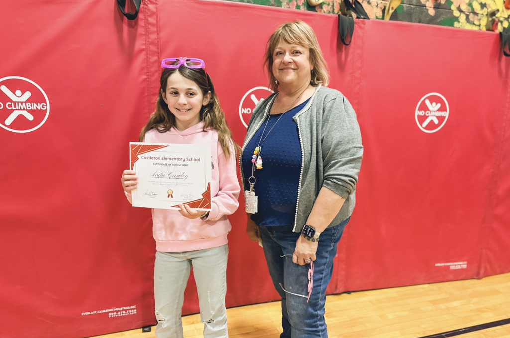 Student holding a Castleton Elementary School certificate stands beside a staff member in the school gym in front of red wall mats.