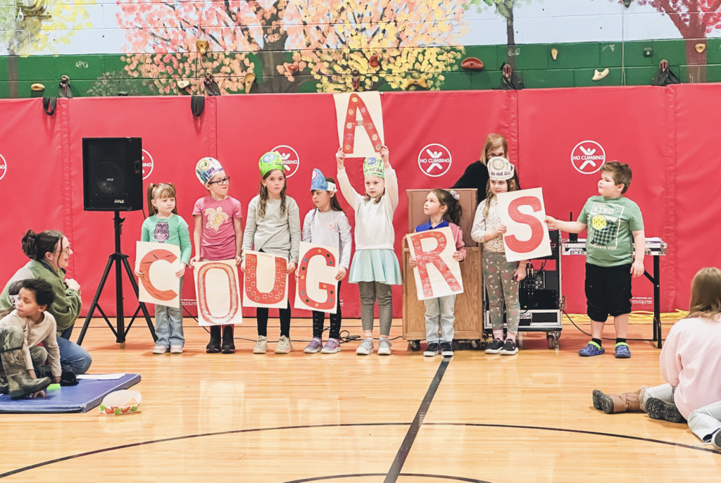 Elementary students wearing themed paper hats hold large letters spelling “COUGARS” during a school assembly in the gym.