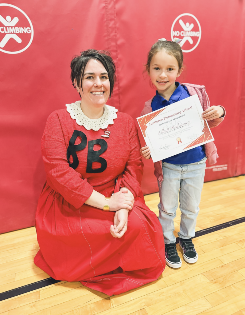 Student holding a Castleton Elementary School certificate stands beside a staff member dressed in a red costume in the gym.