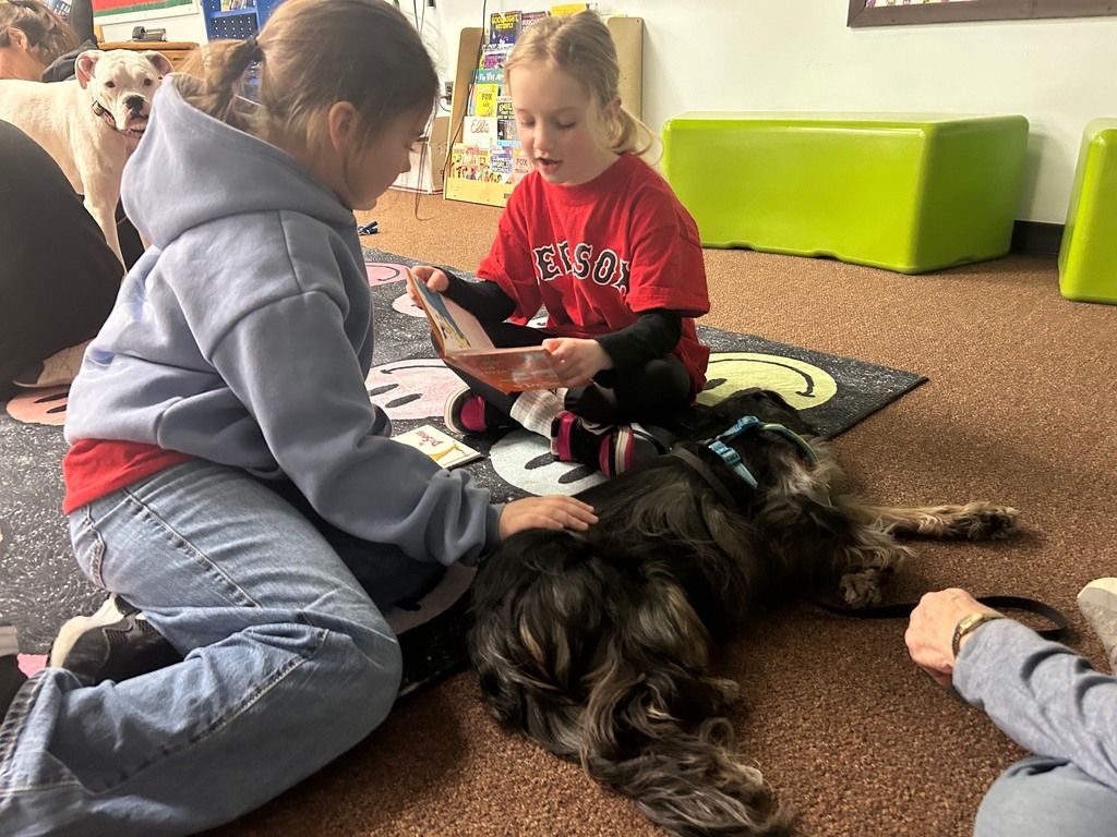 Two students sit on a rug in the school library reading a book aloud while a small therapy dog lies beside them.