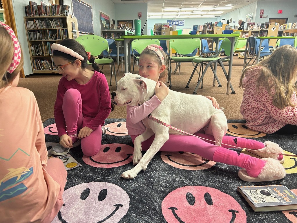Students sit together on a rug in the library while one student gently hugs a white therapy dog during a reading activity.