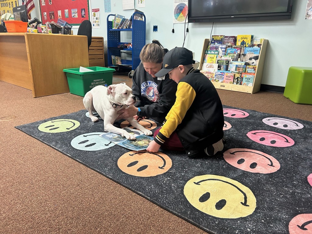 A student and adult sit on a classroom rug reading a book while a white therapy dog lies calmly next to them.