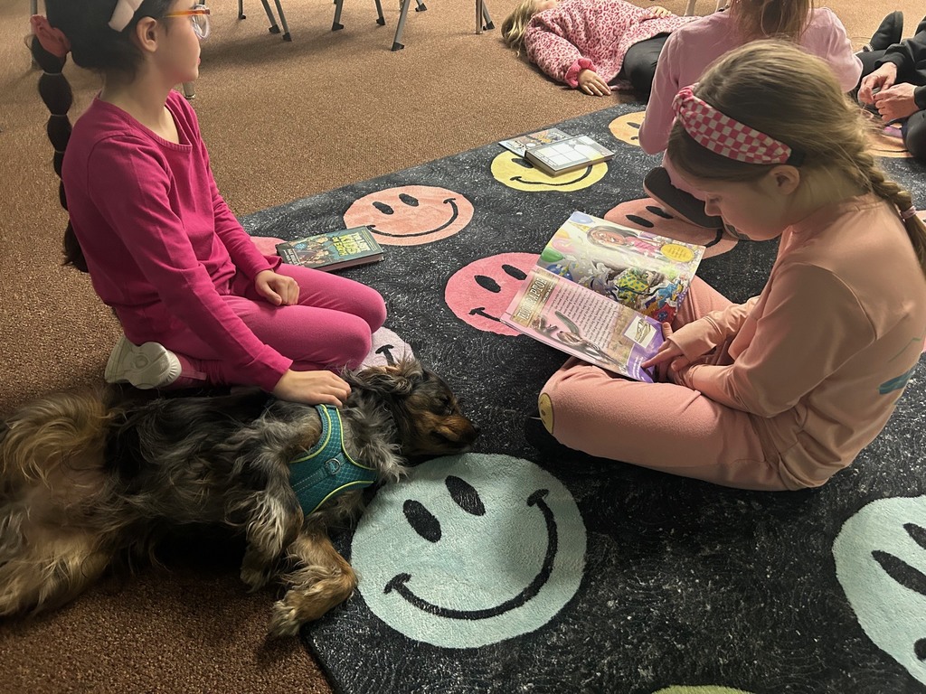 Two students sit on a rug reading books while a small therapy dog rests beside them.