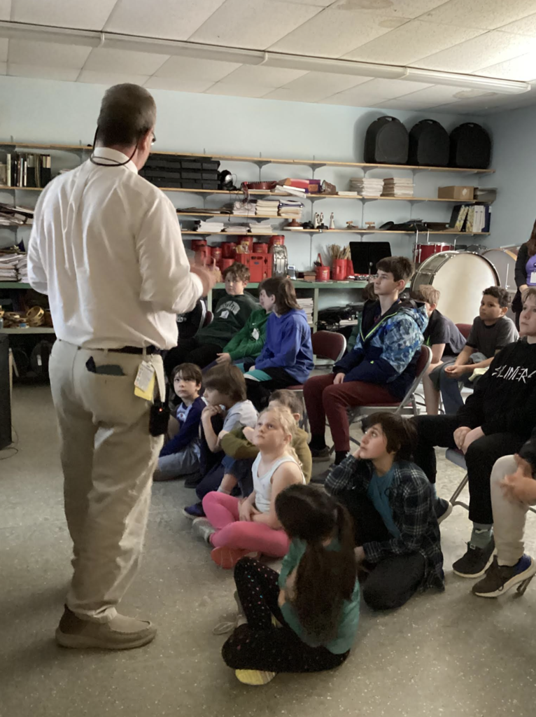 A teacher stands at the front of a music classroom speaking to a group of students seated on the floor and in chairs while shelves of instruments and supplies line the wall behind them.