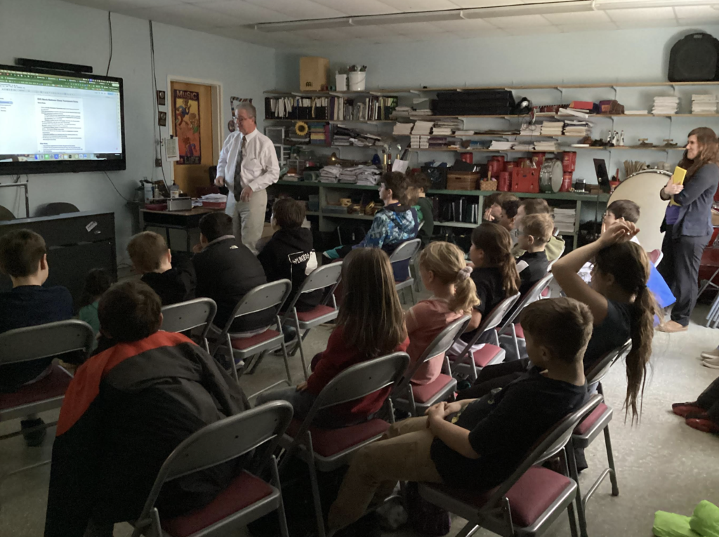 Students sit in rows of folding chairs watching a teacher speak beside a large screen displaying a document in a music classroom.