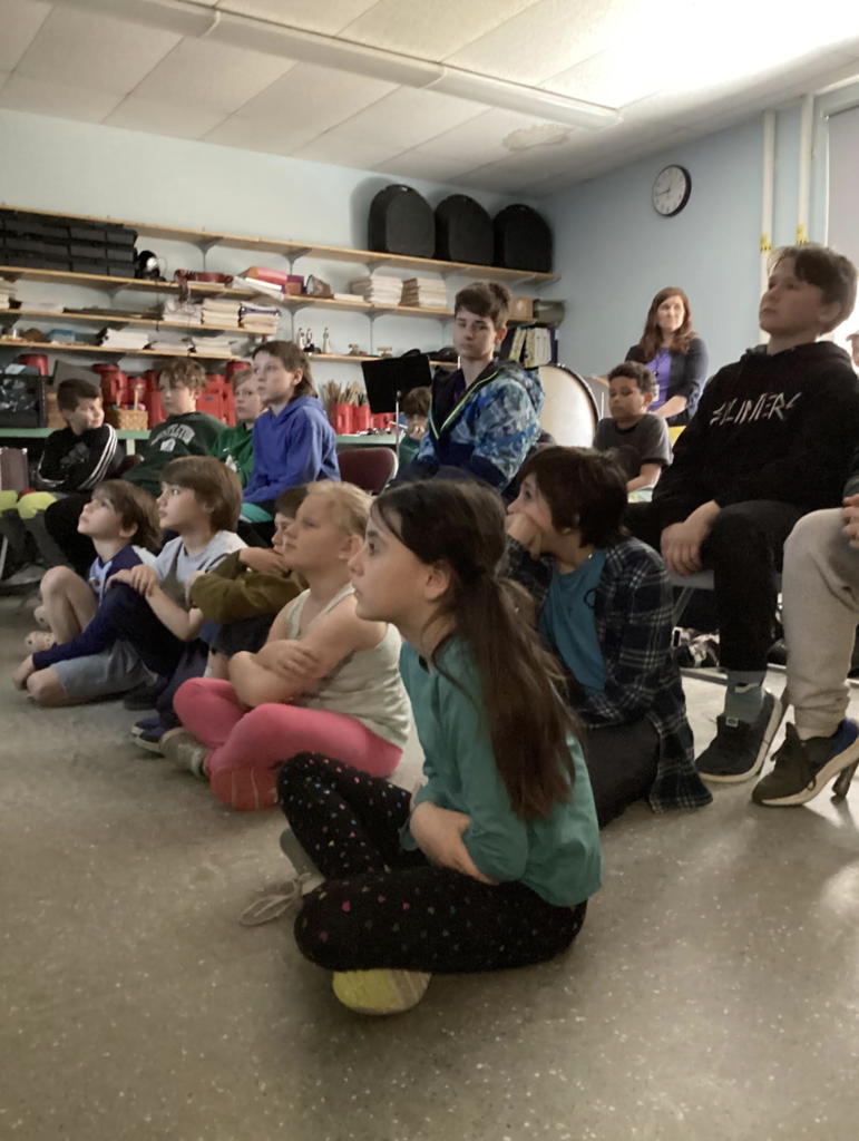 Students sit on the floor and in chairs listening attentively during a classroom presentation while a teacher stands nearby.