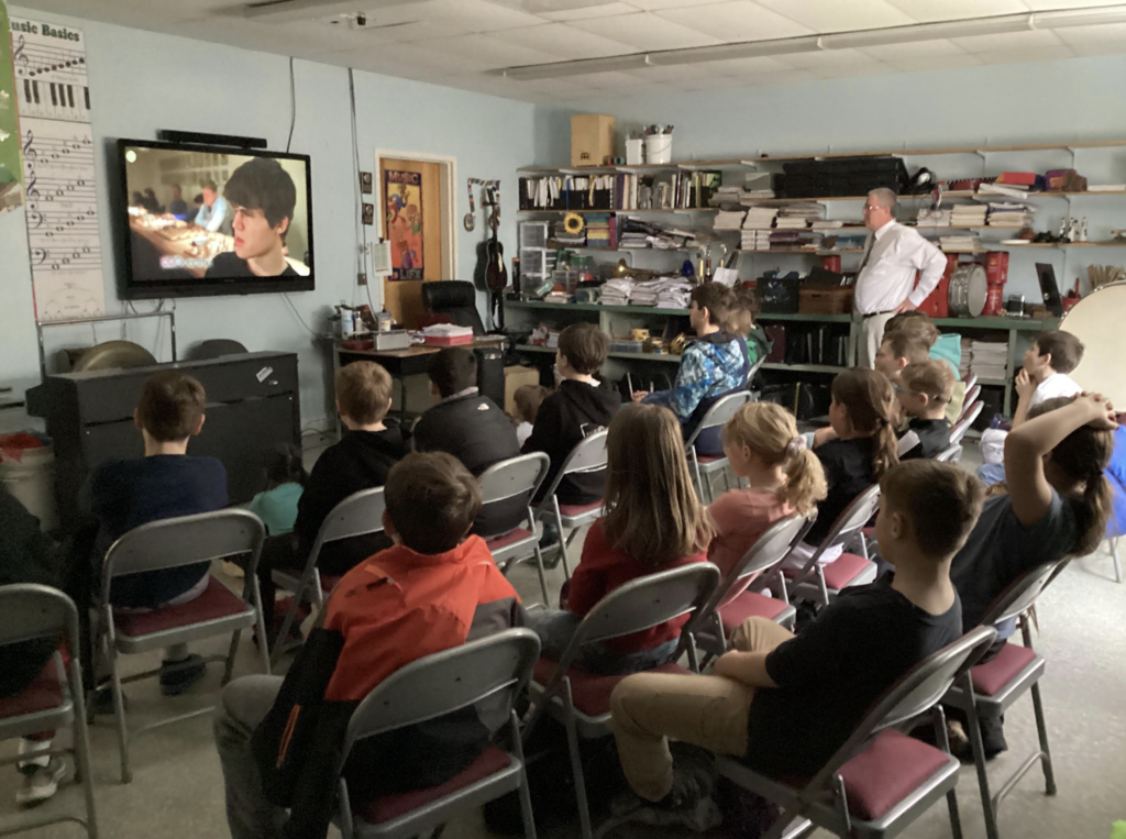 Students sit in rows watching a video on a large screen at the front of the classroom while a teacher stands to the side.