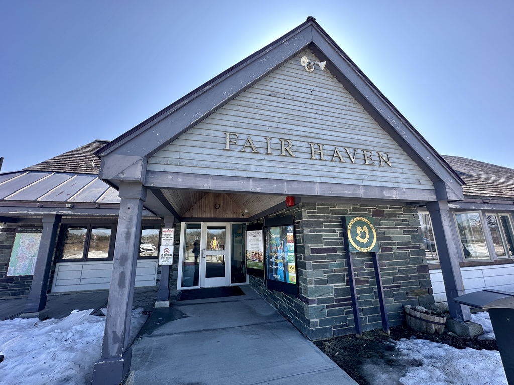 The entrance of the 'FAIR HAVEN' welcome center, built with light-colored wood siding and grey slate-stone, featuring a large gabled entryway and a green state seal sign on a sunny day.