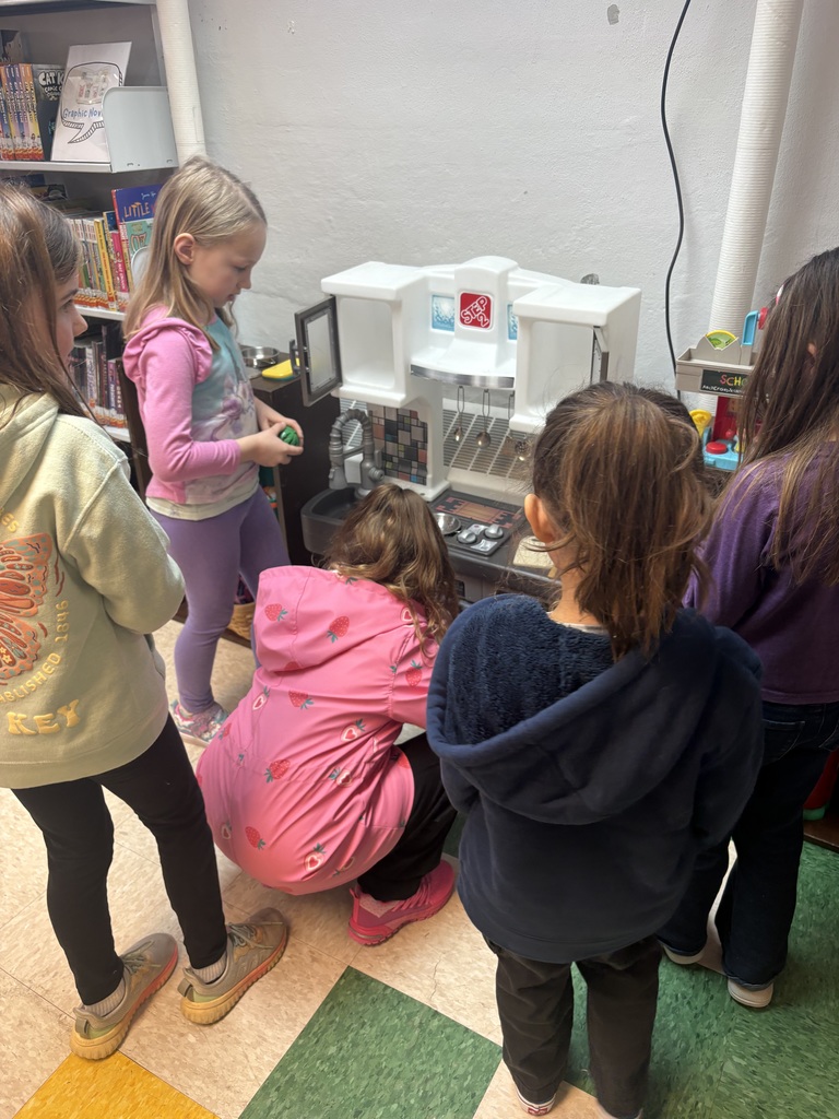 A small group of young girls gathers around a white toy play kitchen, collaborating and exploring the play set together.