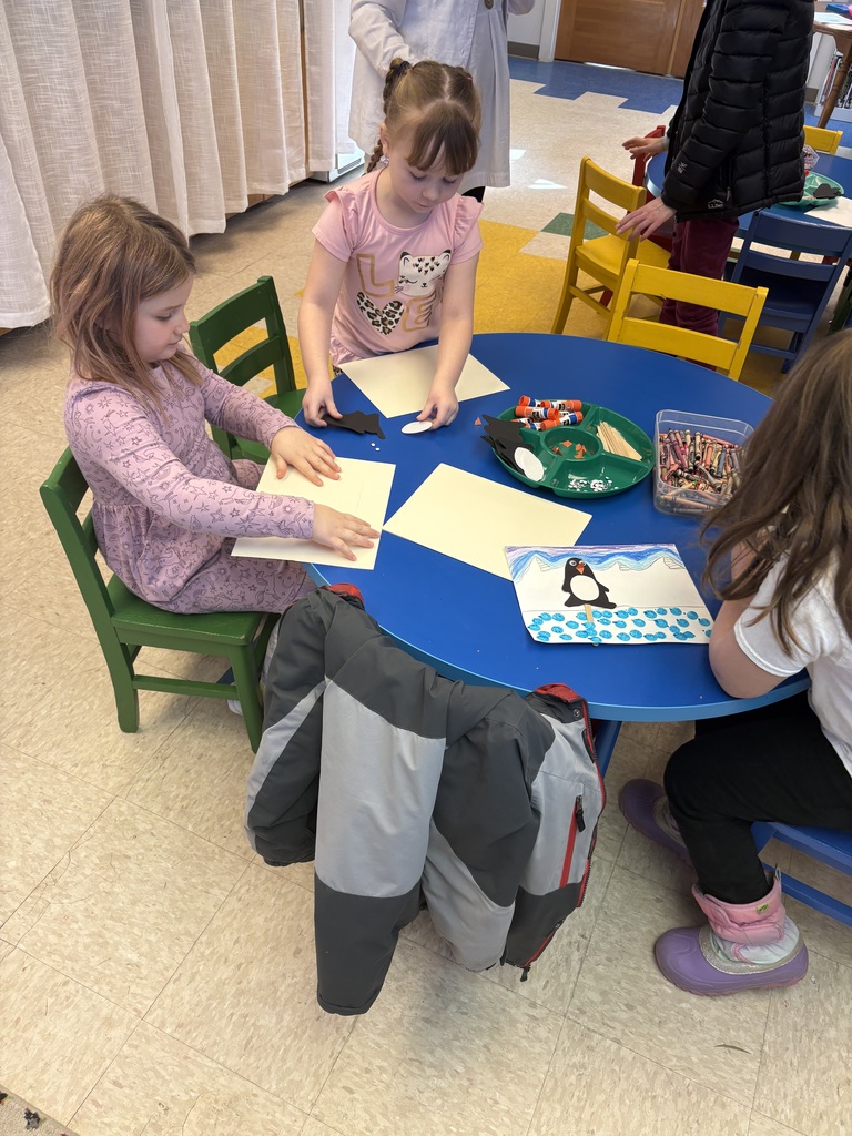 Two first-grade girls are seated at a round blue table, actively working on individual penguin crafts with paper, glue, and crayons.