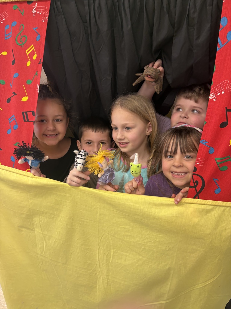 Five cheerful young children smile and peek out from behind a colorful puppet theater, displaying their finger puppets.