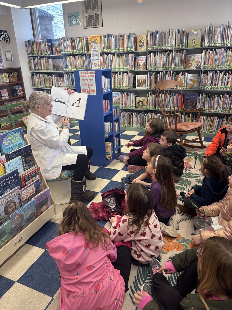 An adult sits and reads a large picture book aloud to a group of first-grade students sitting attentively on a rug in a library setting.
