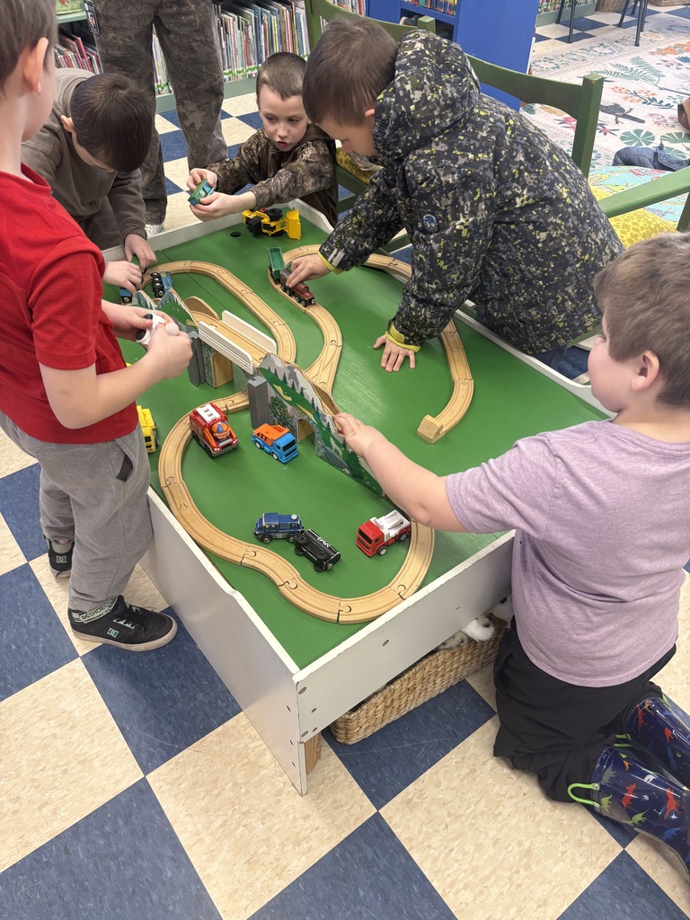 Four young boys actively play with wooden trains and cars around a dedicated train table in a children’s area.