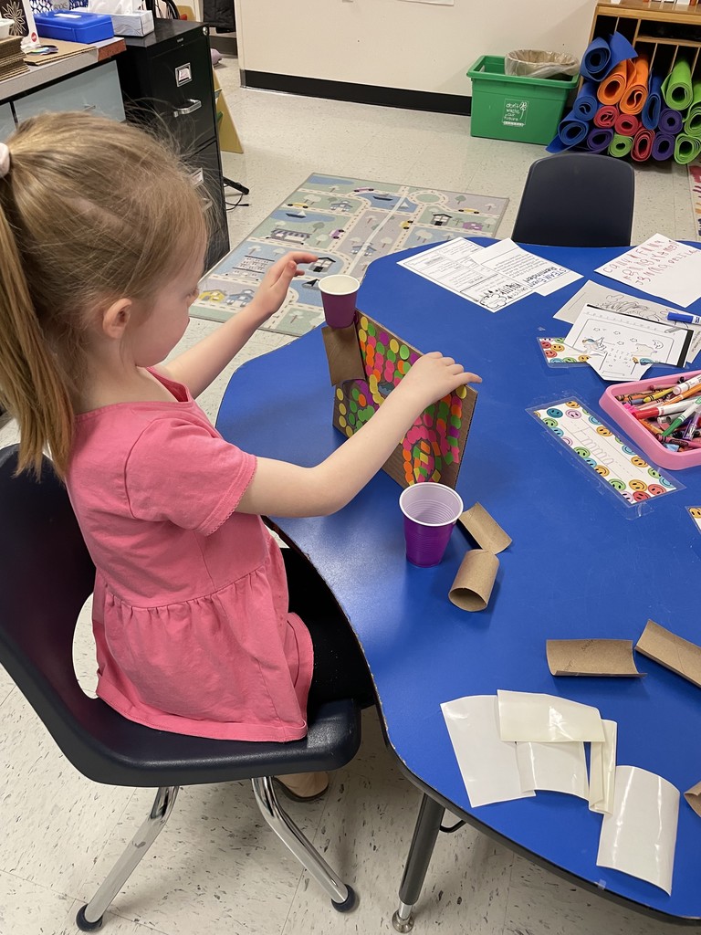 A young girl in a pink dress is working at a blue kidney-shaped table. She is carefully adjusting a cardboard marble run that features a purple cup at the top and a cardboard tube ramp. Several more cardboard tube segments and white adhesive strips are scattered on the table in front of her.  