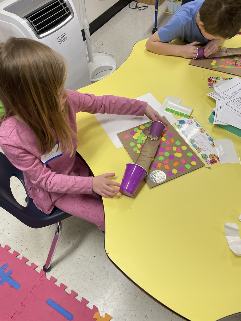 A young girl in a pink patterned outfit sits at a yellow table, carefully aligning a cardboard tube that connects a purple cup at the top to another cup at the bottom of her project. Her cardboard base is heavily decorated with neon orange, green, and pink square and circle stickers.