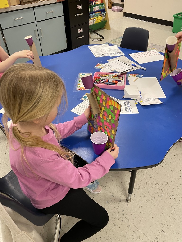 A young girl with blonde hair in a ponytail and a pink long-sleeved shirt sits at a blue classroom table. She is holding a cardboard sheet vertically, which is decorated with colorful neon dot stickers. A purple plastic cup is taped to the top of the cardboard, and she is positioning another purple cup at the bottom to catch a marble.  