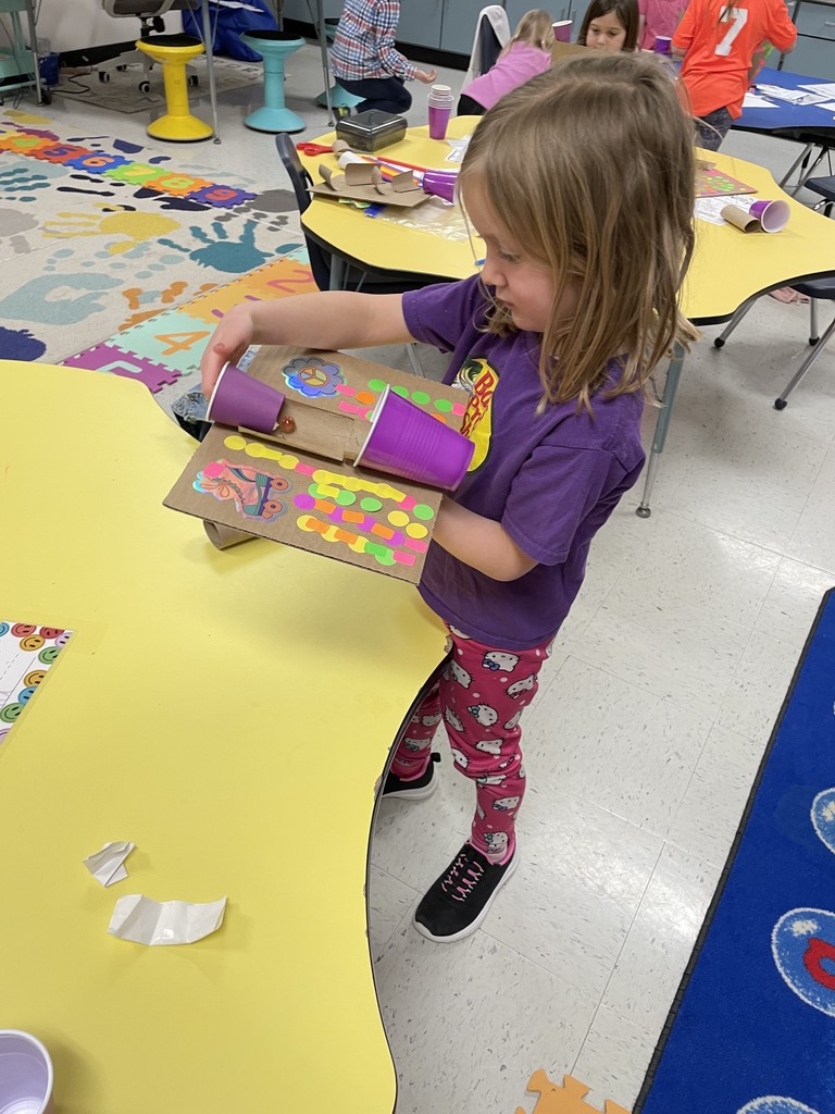 A young girl wearing a purple shirt and Hello Kitty leggings stands at a yellow table, holding her finished marble run. The project features two purple cups and a cardboard track. She is looking down at a small marble resting in the track, testing the gravity-fed design.
