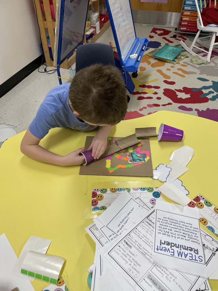  A high-angle shot of a boy in a blue t-shirt leaning over a yellow table. He is using both hands to attach a purple cup and a cardboard tube to his decorated cardboard base. A "STEAM Event Reminder" flyer is visible on the table amongst various crafting scraps.  