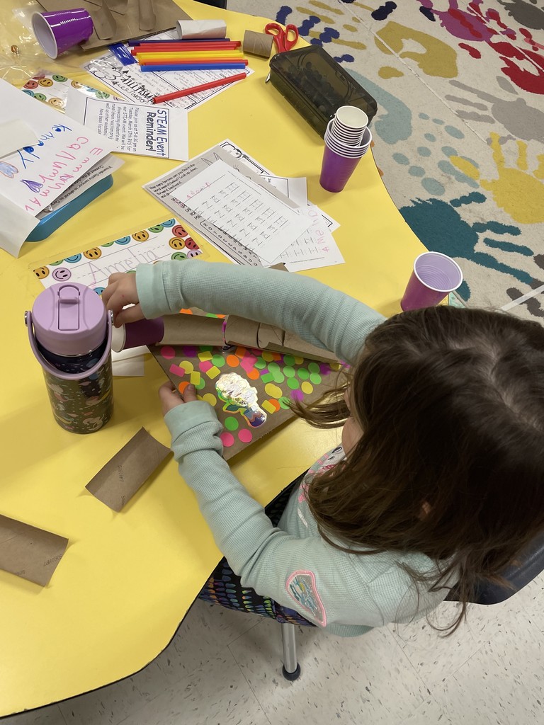  An overhead shot of a girl in a light green textured shirt working at a yellow table. She is assembly a marble run on a cardboard base decorated with bright stickers and a large holographic sticker. Her table is filled with supplies, including purple cups, cardboard tubes, a colorful water bottle, and worksheets.  