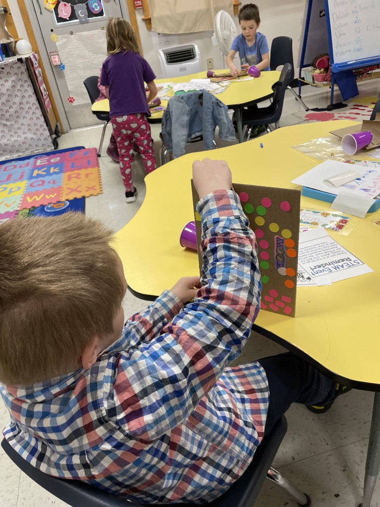  A young boy in a blue and red plaid shirt sits at a yellow table, viewed from behind. He is holding his cardboard marble run upright, focusing on the placement of his neon-colored stickers and tracks. In the background, other children are visible working on similar projects in a brightly decorated classroom. 