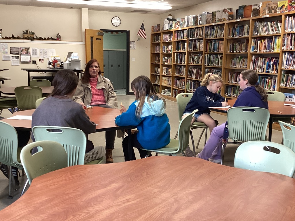 A candid photograph taken inside a well-stocked school library. In the middle ground, an adult woman with reddish-brown hair, wearing a beige jacket and pink top, is seated at a large, rounded wooden table with four young girls. She appears to be speaking or interacting with them. The girls are engaged in writing or drawing on papers in front of them. One girl in the center wears a distinctive bright blue and teal hooded jacket, and another on the right wears a navy sweatshirt.  They are seated on mint-green plastic chairs. The background is dominated by tall wooden bookshelves packed tightly with books. On the back wall, there is an American flag, a wall clock, a bulletin board with posters, and a doorway leading into another room. In the foreground, another unoccupied wooden table and several other green and white chairs are visible. The lighting is warm from overhead fluorescent lights.
