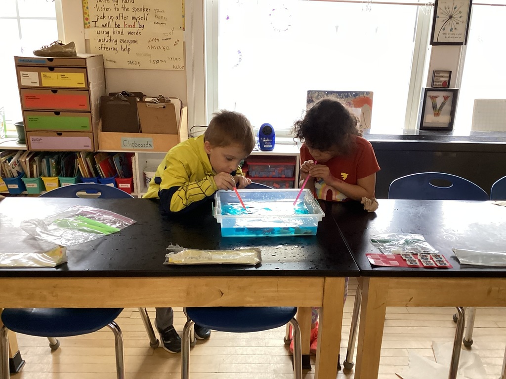 Two children blow through straws into a container of blue water on a table in a sunlit classroom.