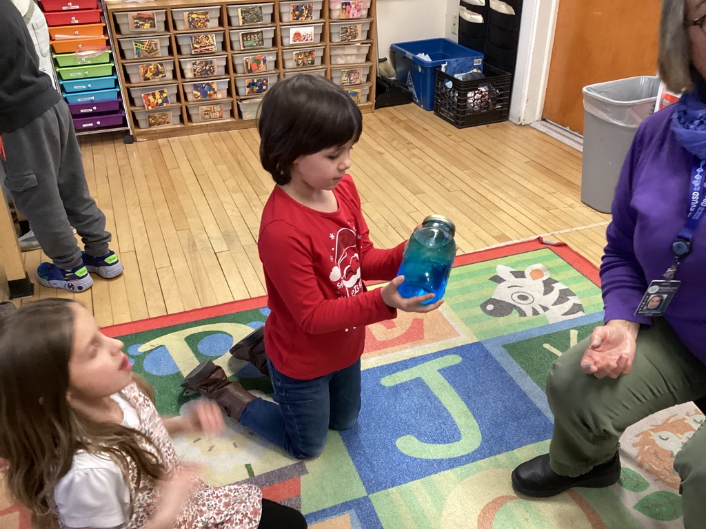 Two girls look up at an adult while one holds a jar of blue liquid on a colorful classroom rug.