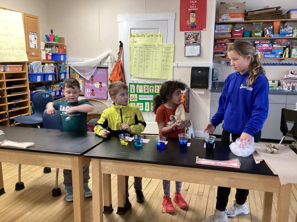 Four children stand behind a table with cups of colored water during a classroom science activity.