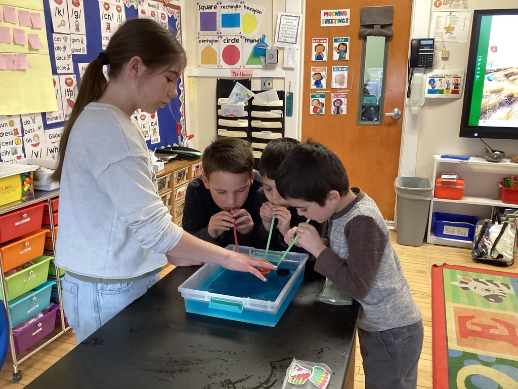 A girl watches as three boys blow bubbles with straws into a bin of blue water on a classroom table.