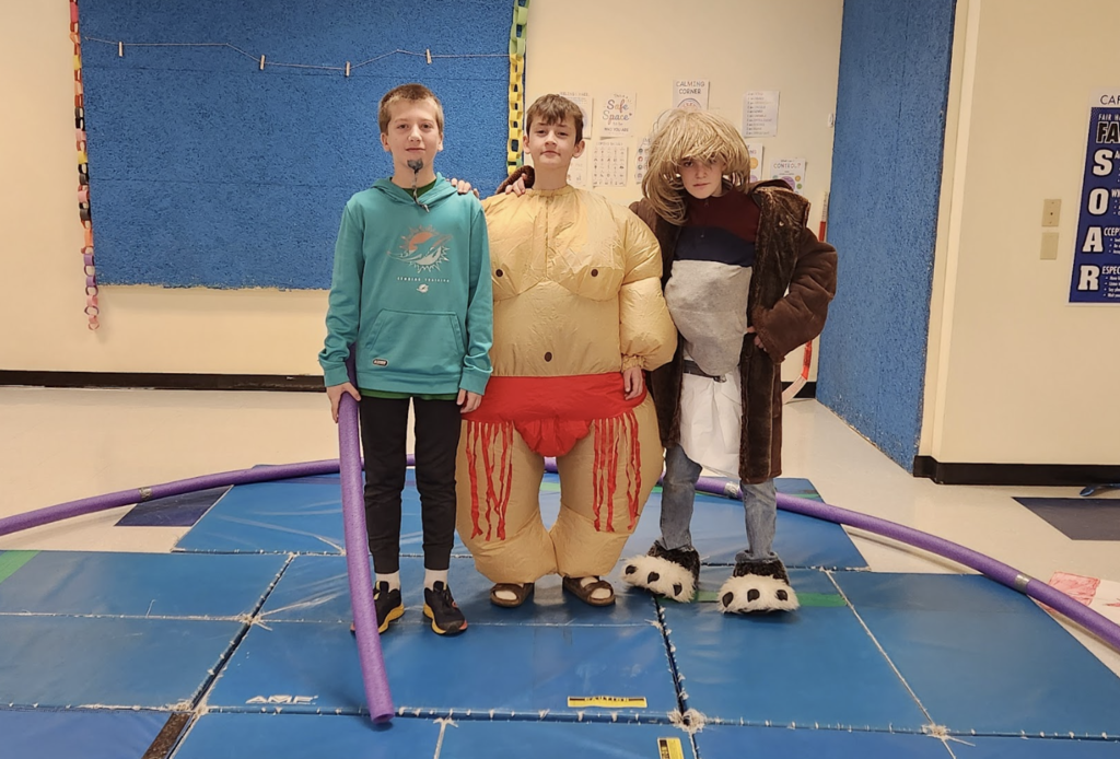 Three students pose on gym mats wearing costumes including an inflatable sumo suit and a large coat with costume accessories.