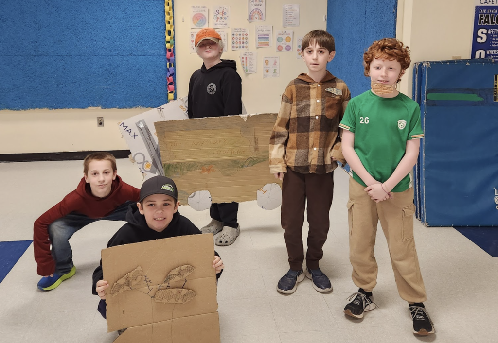 A group of students pose with handmade cardboard props, including a cardboard vehicle and other creative costume pieces for their air band performance.
