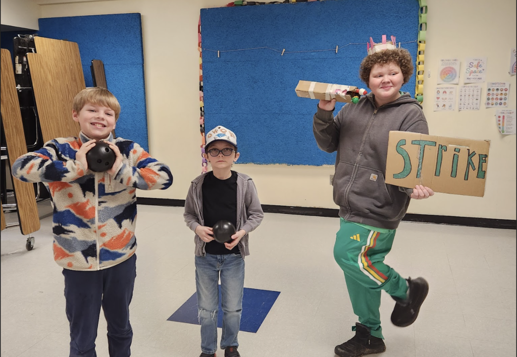 Three students pose with props including small bowling balls and a cardboard sign that reads “Strike,” as part of a creative air band act.