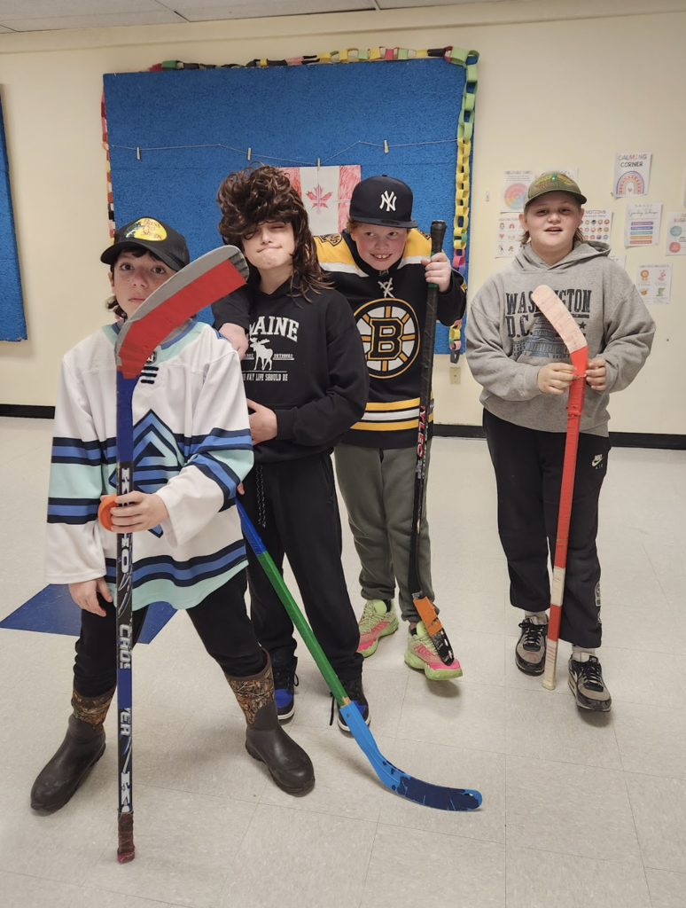 Four students hold hockey sticks and pose like a team during their air band performance, wearing sports jerseys and themed outfits.