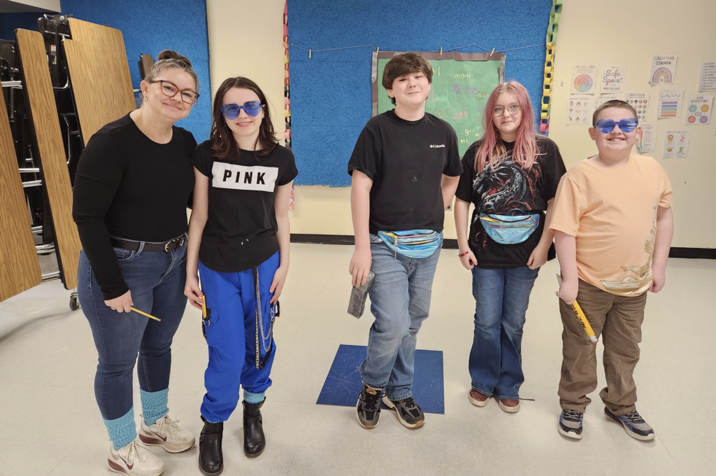 A teacher stands with several students wearing sunglasses and casual outfits as part of their air band group presentation.