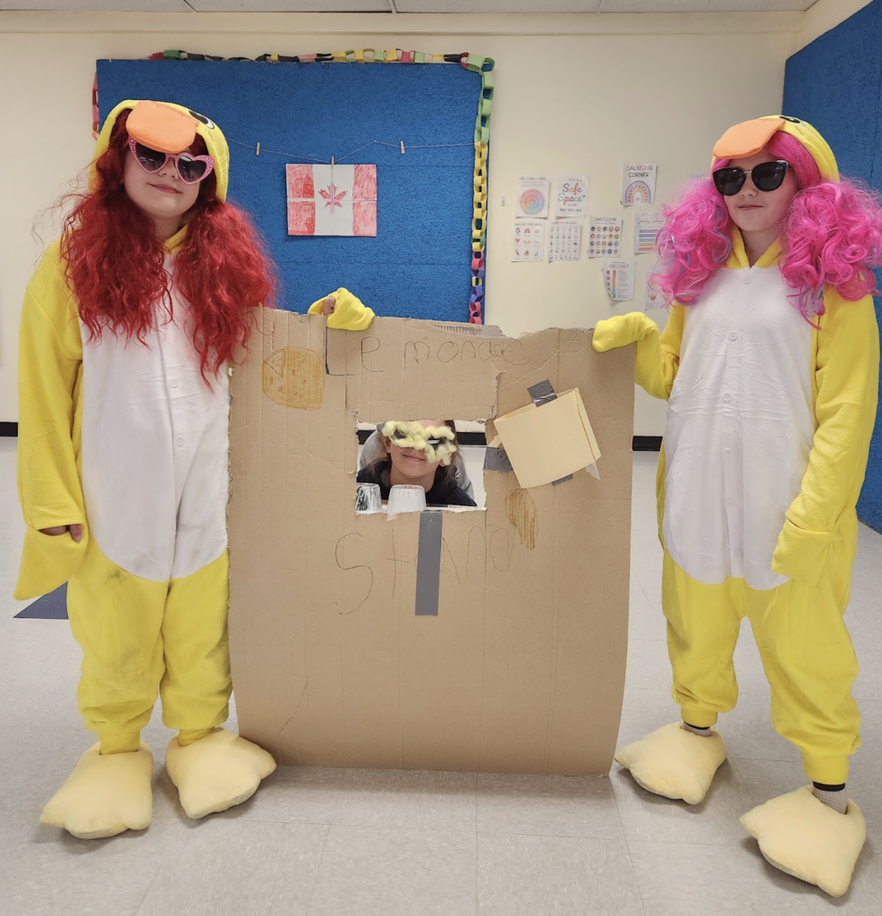 Two students wearing yellow duck costumes and colorful wigs stand beside a cardboard prop stage during their air band performance.
