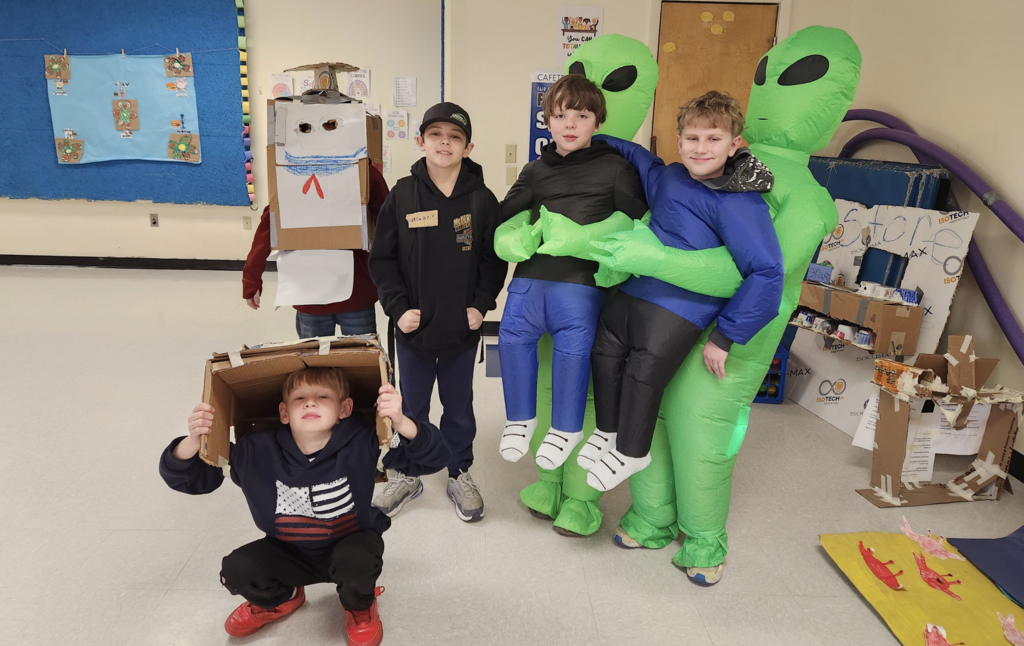 A group of students in costumes, including inflatable alien outfits and cardboard creations, pose together during an air band team activity.
