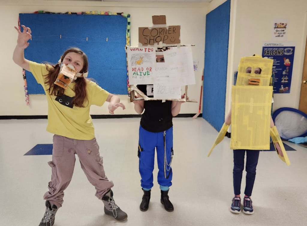 Three students pose with cardboard costumes and props, including a cardboard shop sign and a bright yellow box costume.