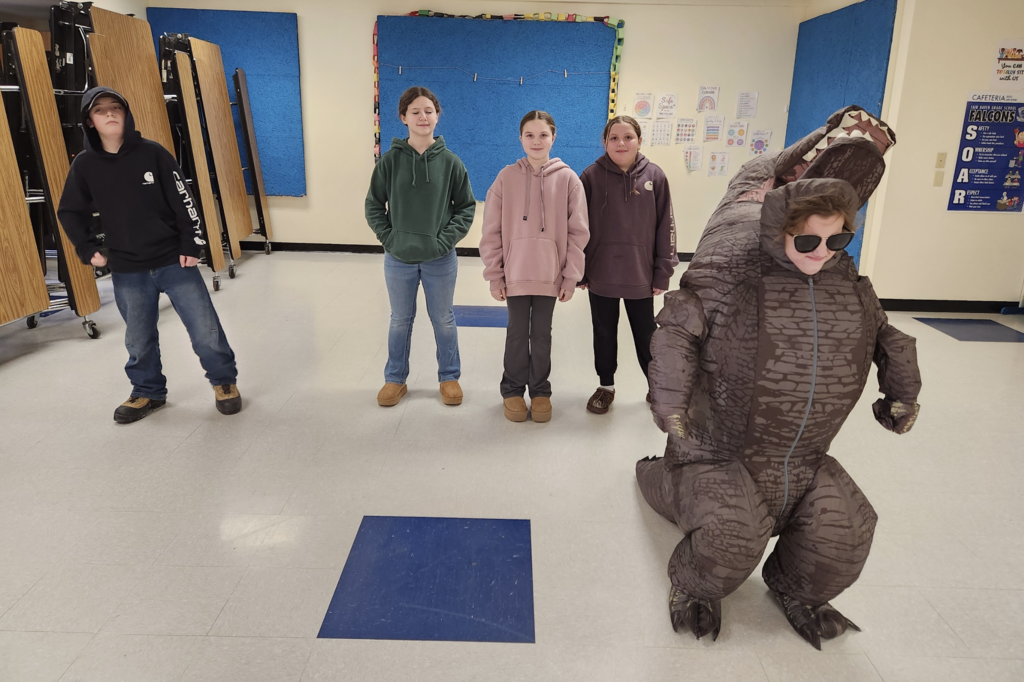A student wearing an inflatable dinosaur costume performs in front of classmates during the air band activity.