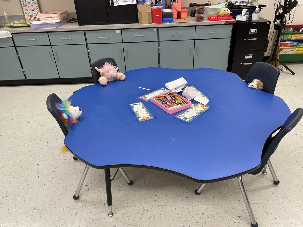  A bright blue, clover-shaped table in a classroom. Three stuffed animals are "sitting" in the chairs: a rainbow-maned lion on the left, a light pink Highland cow at the top, and a small brown and white animal on the right. In the center of the table, there is a pink tray filled with crayons and several laminated name tags. In the background, there is a row of light blue cabinets and a black filing cabinet.